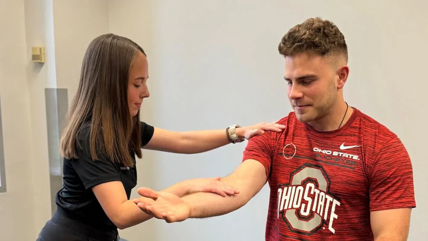 A student practicing physical therapy on a participant wearing an Ohio State jersey, gently touching their shoulder and elbow.