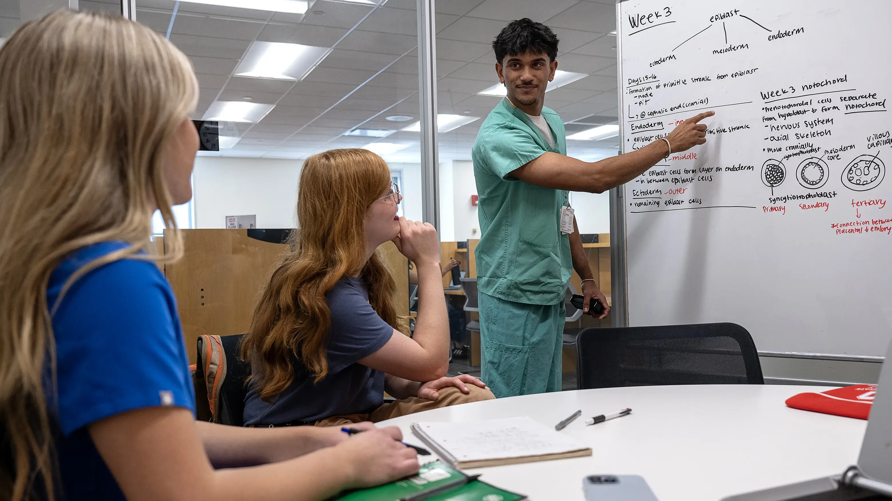 A standing student points to information on a white board while two other seated students watch