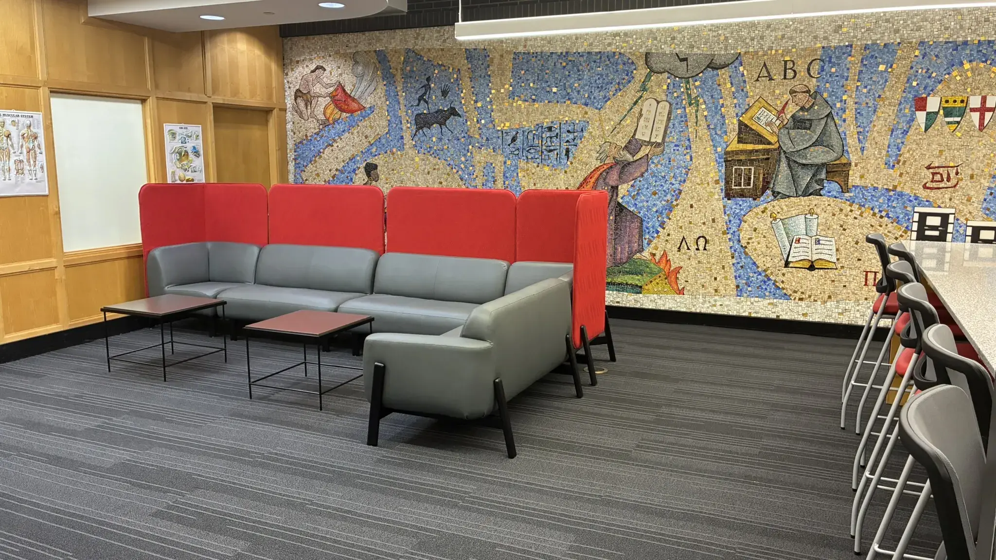 Contemporary lounge featuring a gray sectional sofa, red backrest panels, and small tables, with a vibrant mosaic wall showcasing educational themes.