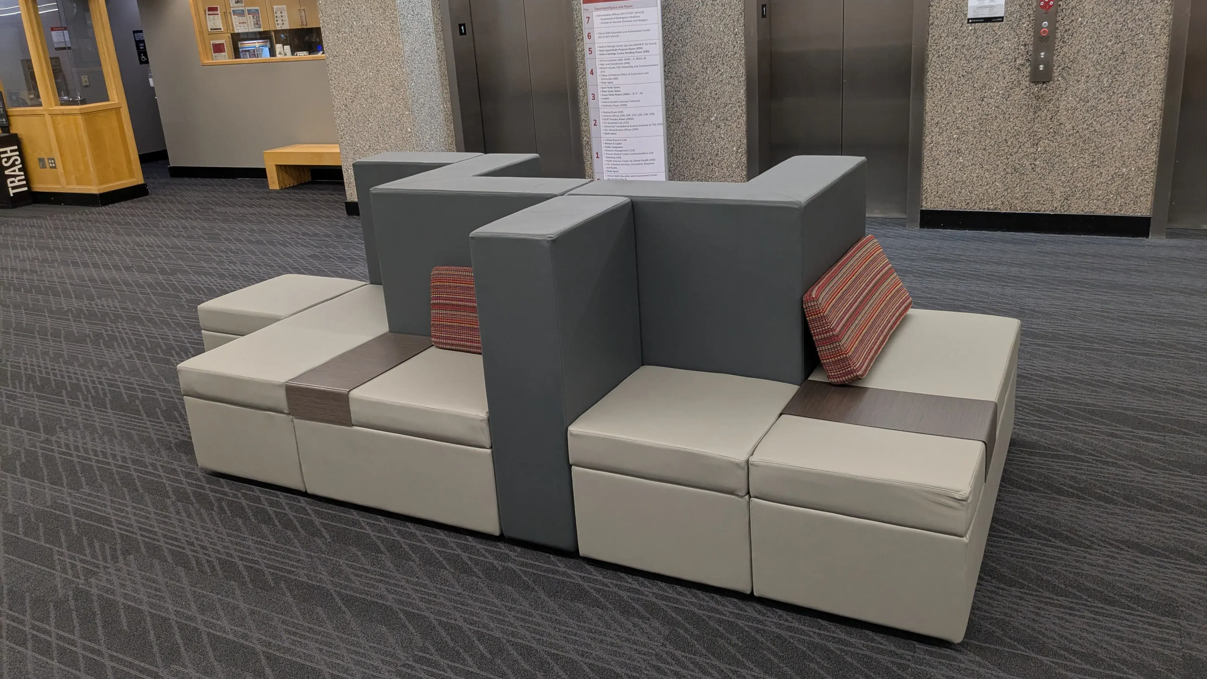 Gray and beige modular seats with red-striped cushions in a lobby, with two elevators and a directory behind. A neat, comfortable setting.