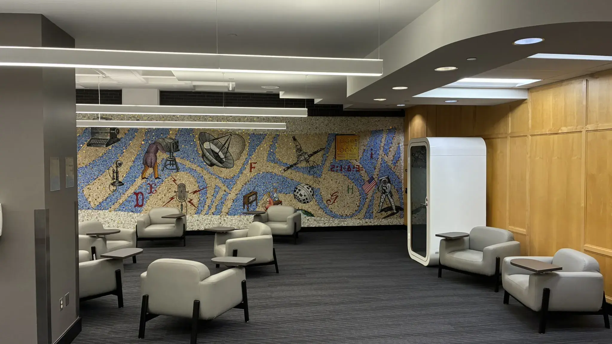 A modern lounge has gray armchairs, small tables on a dark carpet, and a vibrant mosaic mural with scientific designs on the back wall.