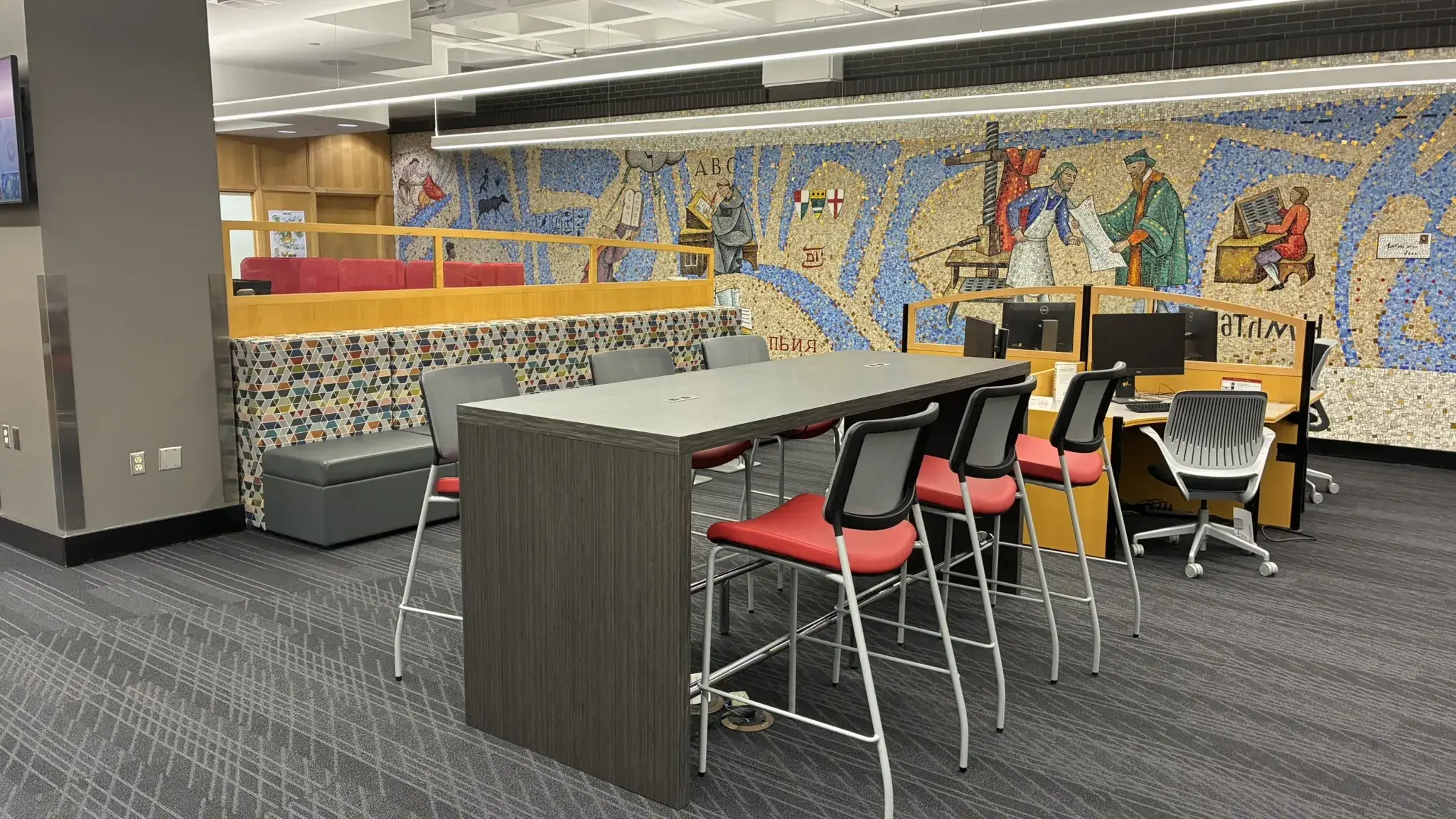 Modern library interior featuring a mosaic mural of historical scenes, a tall table with red chairs, workspaces, and a quiet, focused atmosphere.