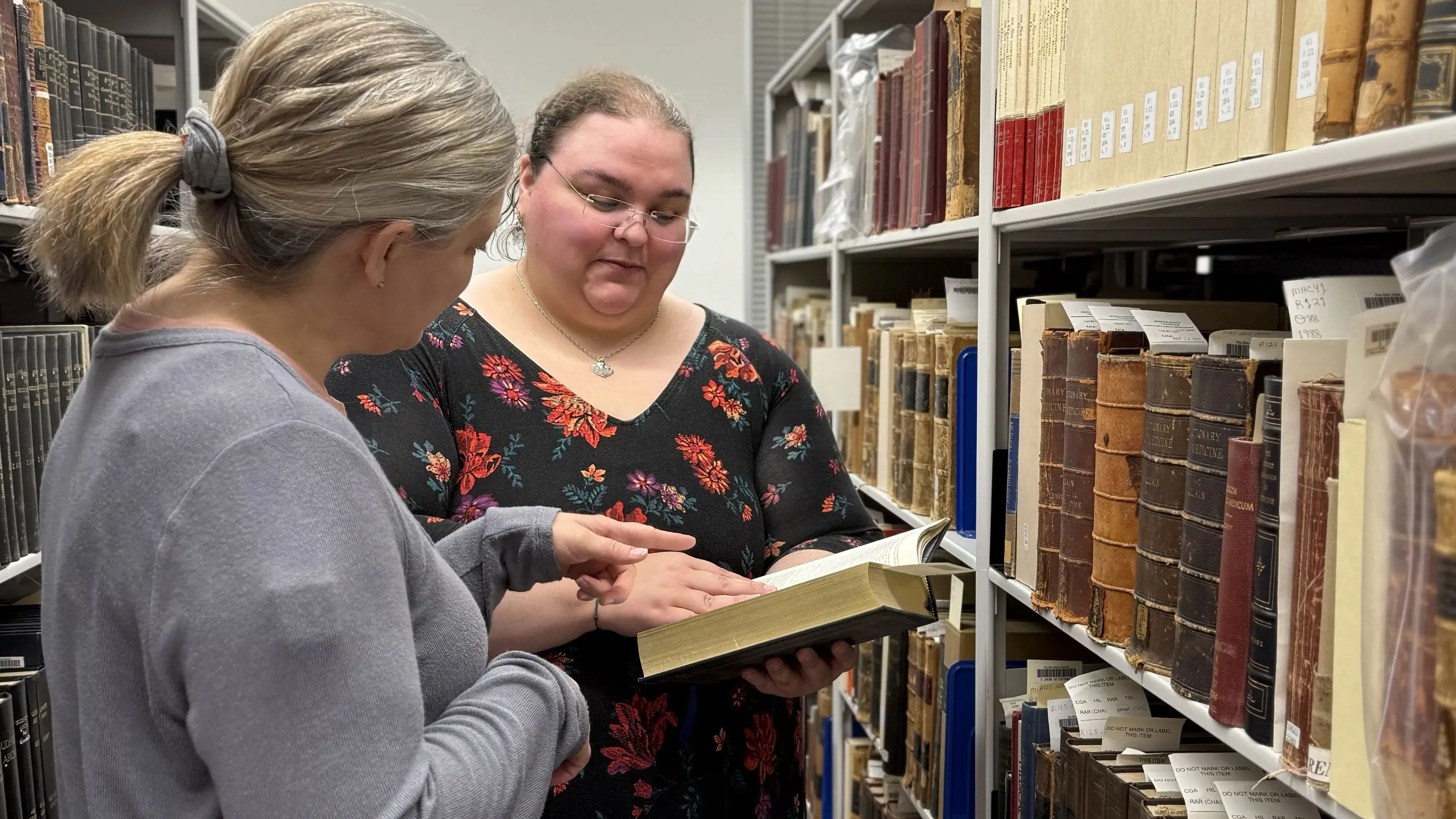 Two female individuals look at a book together in a library book stack