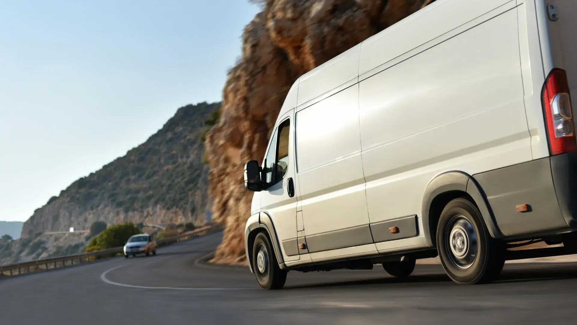 White van on a scenic curved mountain road, passing rocky cliffs. A car follows in the distance. The scene conveys motion and adventure.